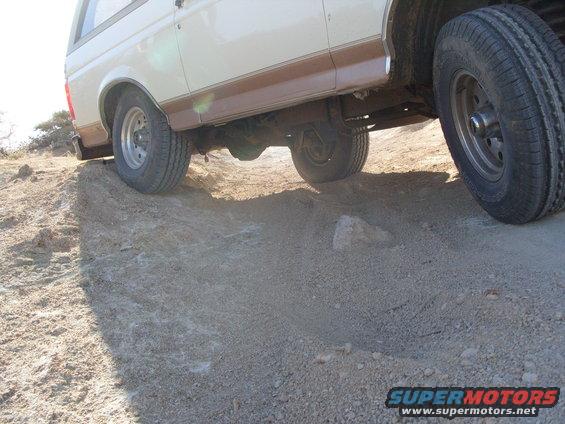 Side view of a white and brown Ford Bronco driving on a dirt hill. The front wheel is turned