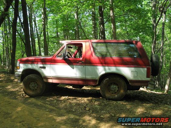 Red and white Ford Bronco with a spare tire mounted on the rear, parked on a dirt path in a forest