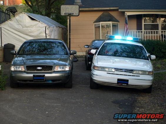 Two Ford Crown Victoria sedans parked in a driveway. One is white with blue lights on top, the other is gray
