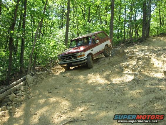 Red and white Ford Bronco driving on a dirt trail in a wooded area