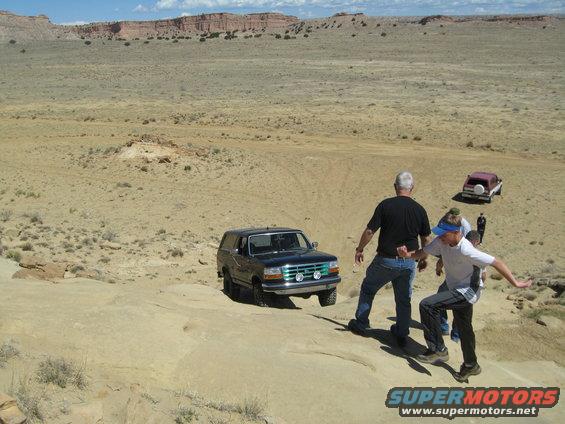 1993 Ford Bronco driving on a rocky desert trail with two people nearby and another vehicle in the background