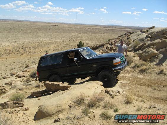 Black Ford Bronco driving over rocks in a desert landscape. Two people are visible