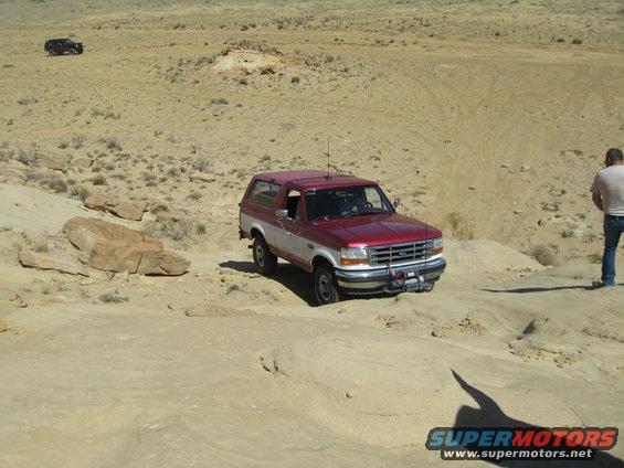 Red and white Ford Bronco driving on a dirt trail with a man standing nearby
