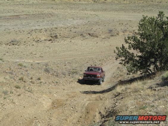 Red pickup truck on a dirt road in a dry, hilly area. The truck appears to be from the early 1990s