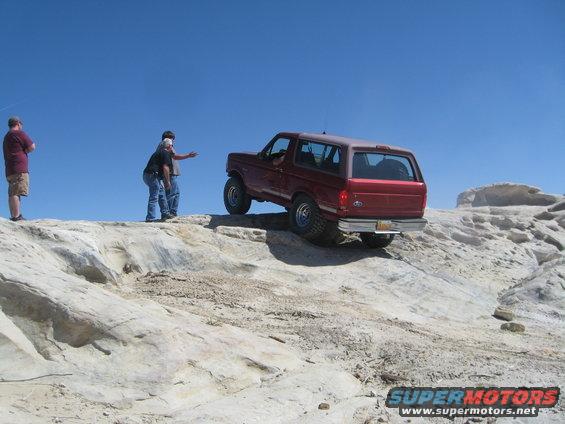 Red 1993 Ford Bronco driving off-road on a rocky hill with two people standing nearby