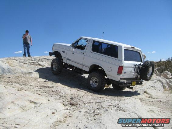 White 1993 Ford Bronco climbing rocky terrain with a person standing on a ridge nearby
