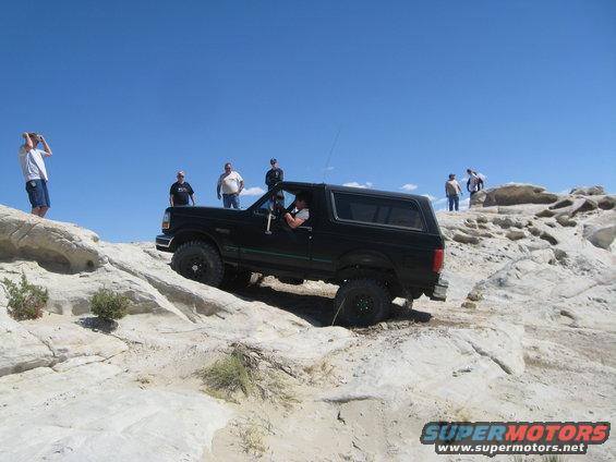 Black 1993 Ford Bronco driving off-road on a bright, sunny day with several people watching