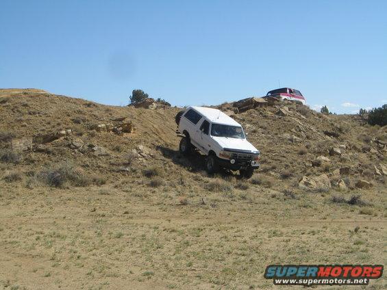 White Ford Bronco driving on a dirt hill with a red and white SUV behind it