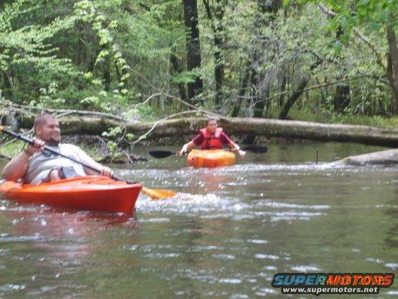 Two people are kayaking down a river surrounded by trees. One person is in the foreground in an orange kayak, and another person is further back in an orange inflatable kayak