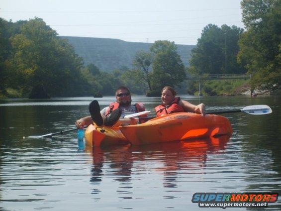 Two people wearing life vests relax in orange kayaks on a calm lake with trees and a dam in the background