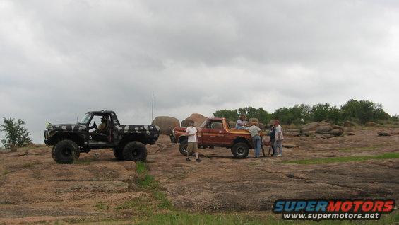 Two modified trucks, one black with camouflage, one orange, parked on a rocky landscape under a cloudy sky