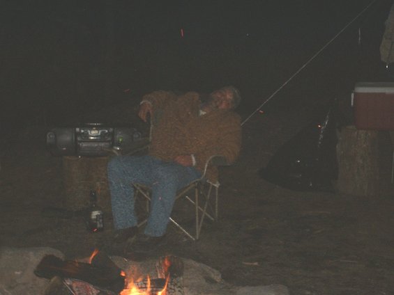 Man in a fuzzy jacket and jeans sits in a folding chair near a campfire, with a boombox on a table behind him