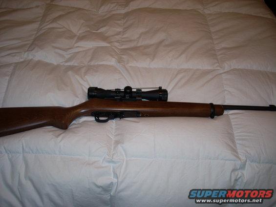 Wooden rifle with a black scope mounted on top, resting on a white comforter