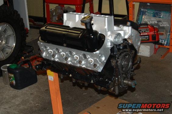 Engine on an orange stand, with a black valve cover and silver intake manifold. A tire and a green gas can are visible to the left