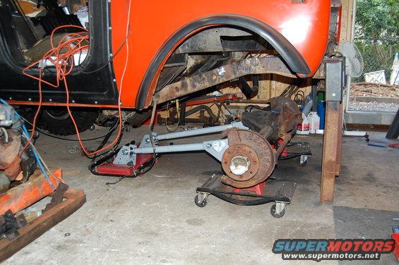 Orange SUV chassis on jack stands, front suspension removed and on dollies. Orange cables run into the open door