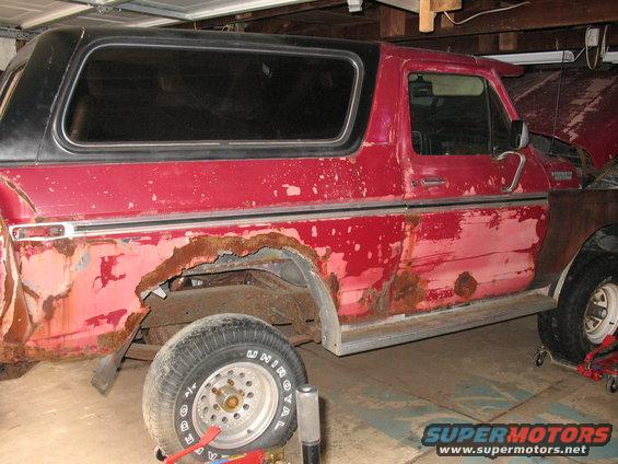 Red Ford Bronco with extensive rust damage on the rear quarter panel and lower body. The vehicle is in a garage with the hood open