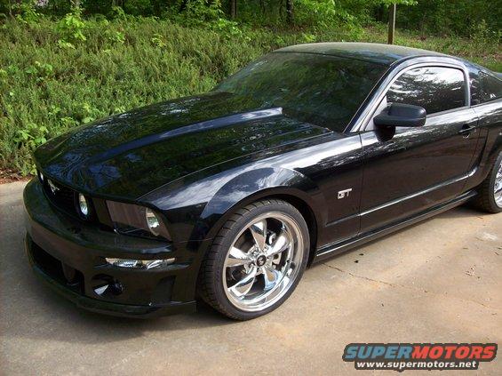 Black Ford Mustang GT parked on concrete with trees behind it. Has a scoop on the hood and chrome wheels