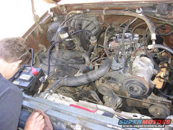 Engine compartment with a man working on the battery; brown vehicle exterior visible