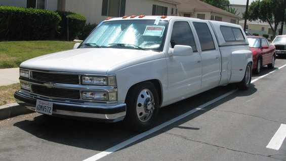 White crew cab dually pickup truck with a camper shell parked on a street. Another red car is parked behind it