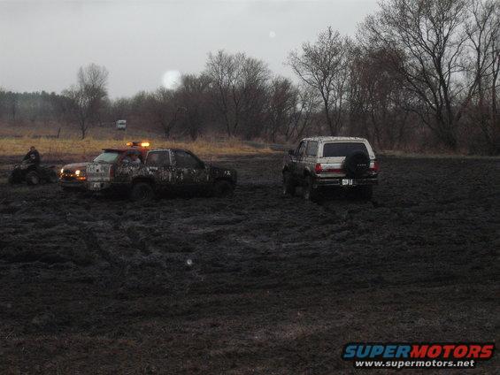 1991 Ford Bronco and a muddy pickup truck stuck in mud, with an ATV nearby
