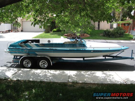 Side view of a teal and white boat on a trailer. Text on hull reads 'Supermotors.net'