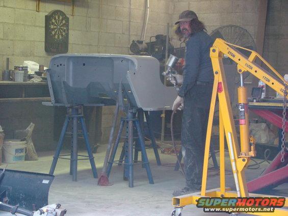 Man sprays primer on Jeep CJ-5 body shell on stands, yellow engine hoist nearby