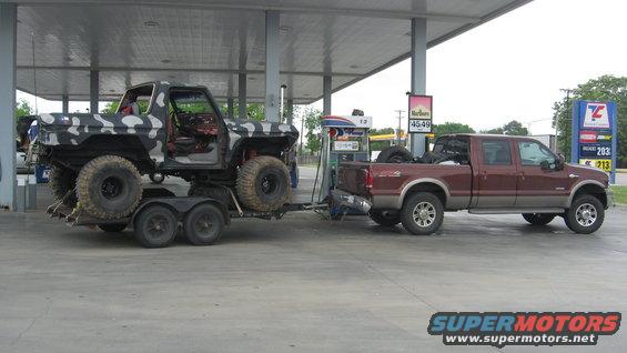 Camouflaged Ford Bronco on trailer being towed by a maroon Ford pickup truck at a gas station