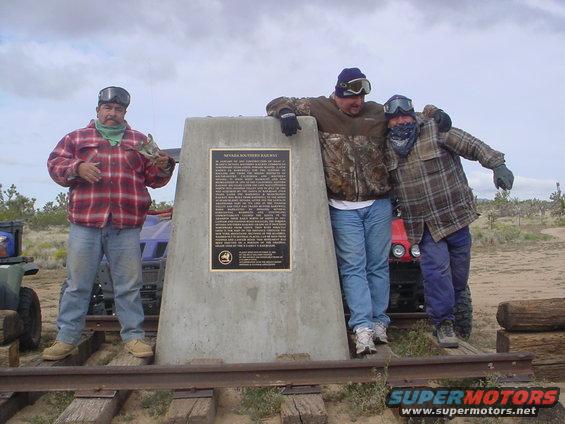 Three men stand by a concrete monument with a historical plaque. ATVs are visible behind them