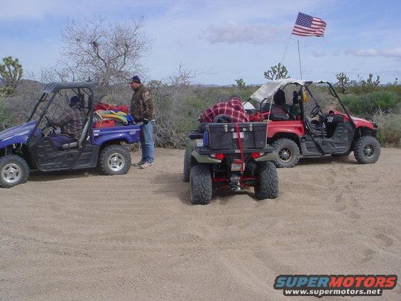 Three ATVs parked on dirt in a desert landscape. One blue, one red, and one green. An American flag flies from the red ATV