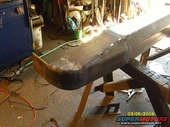 Custom metal bumper being fabricated, supported by wooden sawhorses. Grinding tools and welding marks are visible