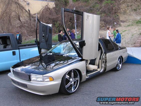 Gold and black sedan with butterfly doors open, chrome rims, and lowered suspension. A blue pickup truck is parked to the left
