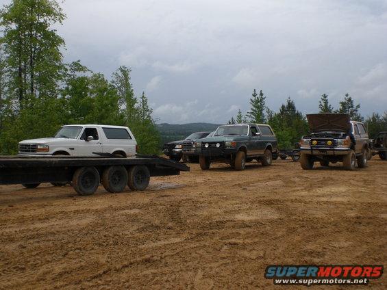 Several Ford Bronco SUVs parked in a muddy outdoor area with trees in the background. One Bronco is on a trailer
