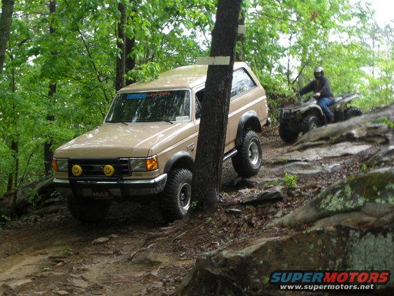 Tan 1989 Ford Bronco navigating a rocky trail, with an ATV rider in the background