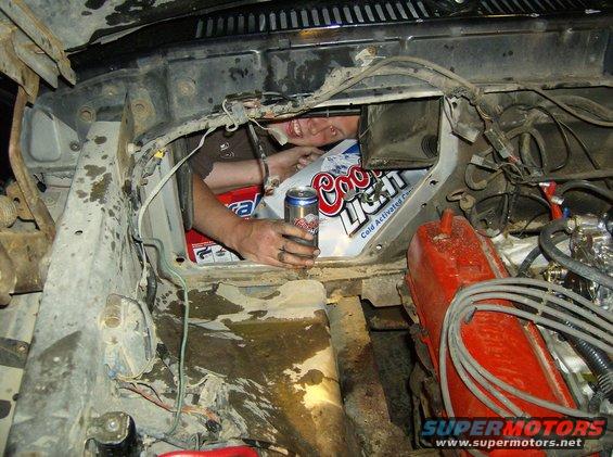 Engine bay with a person reaching in, holding a Coors Light can, with a red valve cover visible