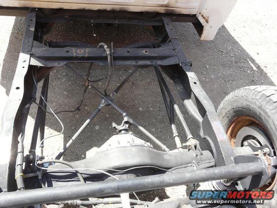 Truck chassis and rear axle, showing leaf springs and driveshaft. White paint on upper body. Tire visible on right