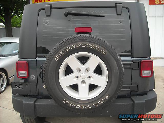 Dark gray Jeep Wrangler rear with spare tire, aftermarket bumper, and a silver car to the left