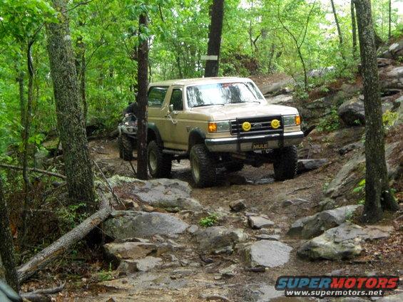 Tan 1989 Ford Bronco navigating a rocky off-road trail with an ATV behind it
