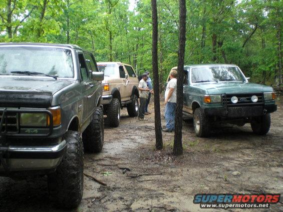 Three lifted trucks parked on a muddy trail in the woods. The green truck in front has a brush guard and auxiliary lights