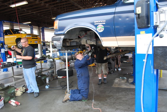 1986 Ford Bronco on a lift with mechanics working underneath. Yellow pickup truck on another lift in background