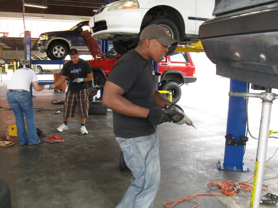 Mechanics work on cars in an auto repair shop. One man uses a drill