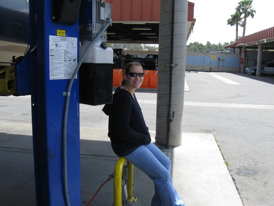 Woman in sunglasses and blue jeans sits on a yellow bar next to a blue car lift. A black car is visible in the background