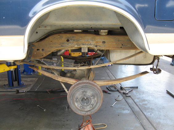 Rear axle with leaf springs and brake drum on jack stands, under a blue and tan vehicle