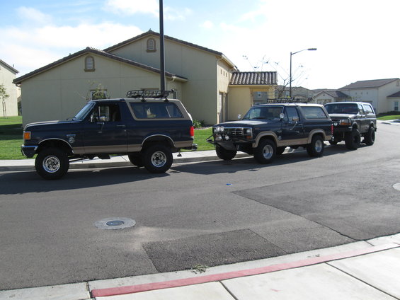 Two dark blue Ford Broncos with tan trim parked on a residential street, one in front of the other