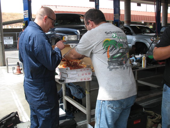 Two men in a garage eating pizza from stacked boxes, with a dark pickup truck and a silver SUV visible in the background
