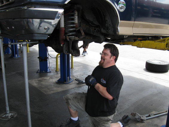 Man in black shirt and shorts kneels under a lifted vehicle, working on the suspension. A tire sits nearby