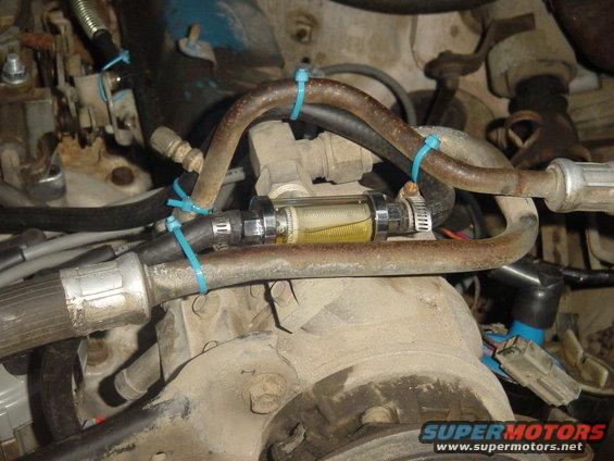 Close-up of a clear inline fuel filter with yellow fluid, connected by hoses and zip ties in an engine bay