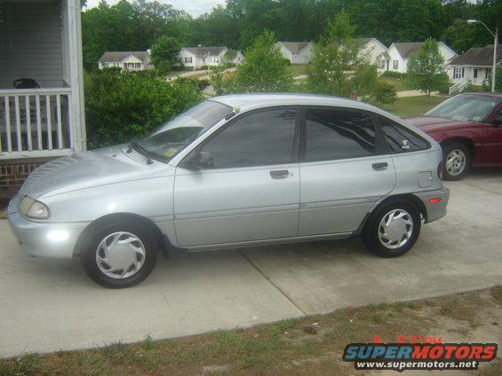 Silver hatchback parked on driveway, red car behind it. Houses and trees in background