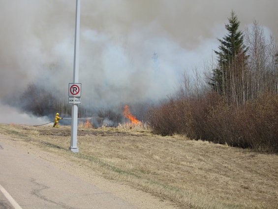 Firefighter in yellow suit sprays water on burning grass with smoke in background. Signpost with 'R' symbol visible