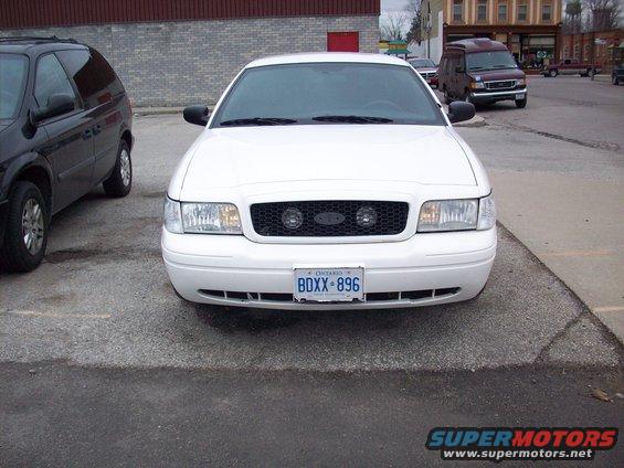 Front view of a white 2003 Ford Crown Victoria with a black grille and Ontario license plate BDXX-896. A black minivan is parked to the left