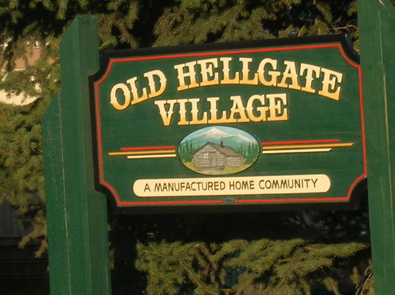 Green sign with yellow lettering reading 'OLD HELLGATE VILLAGE' above a picture of a log cabin and mountains. Below that, it says 'A MANUFACTURED HOME COMMUNITY'